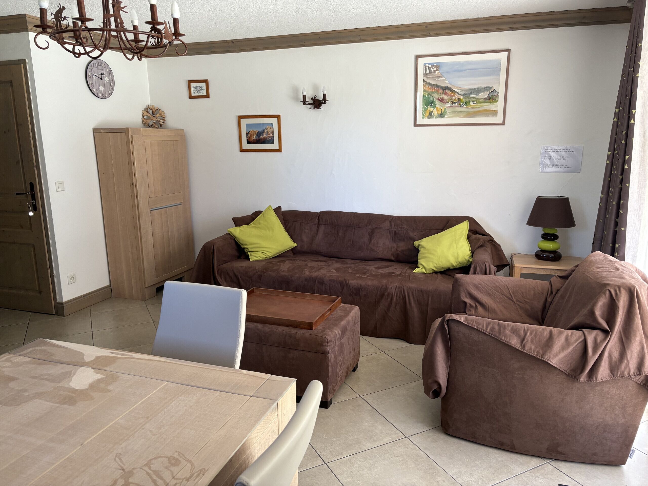 A living room with a brown sofa, two green cushions, a wooden cabinet, a table with a laptop, and a painting on the wall.