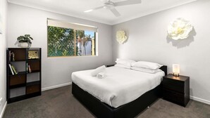 A modern bedroom with a large bed, a ceiling fan, a bedside table with a lamp, a bookshelf, and a window with a view of greenery. - Charming 3-bed Apartment Near Local Shops (Brisbane)