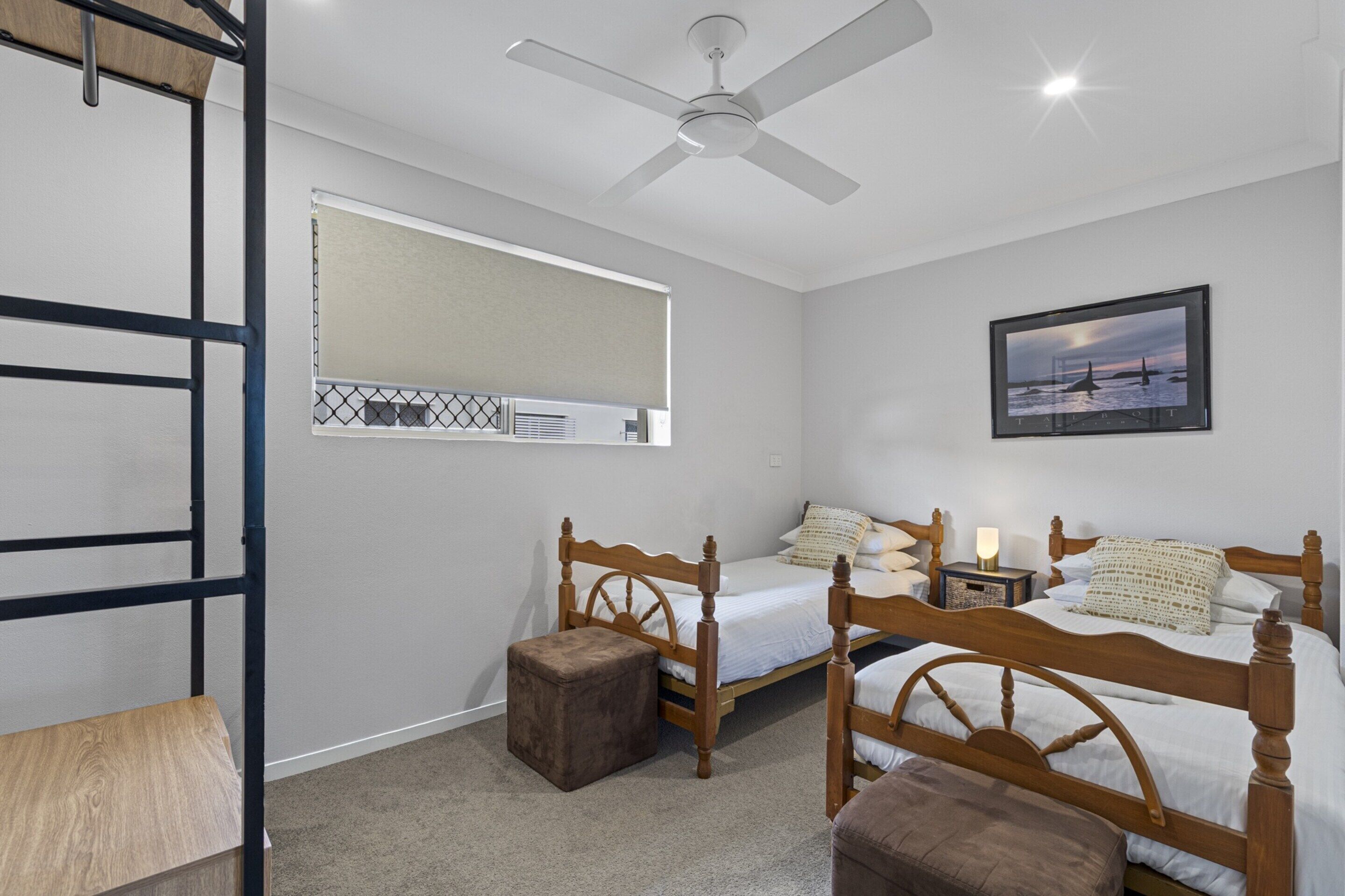 A bedroom with two wooden beds, a ceiling fan, a window with blinds, a nightstand with a lamp, and a framed picture on the wall.