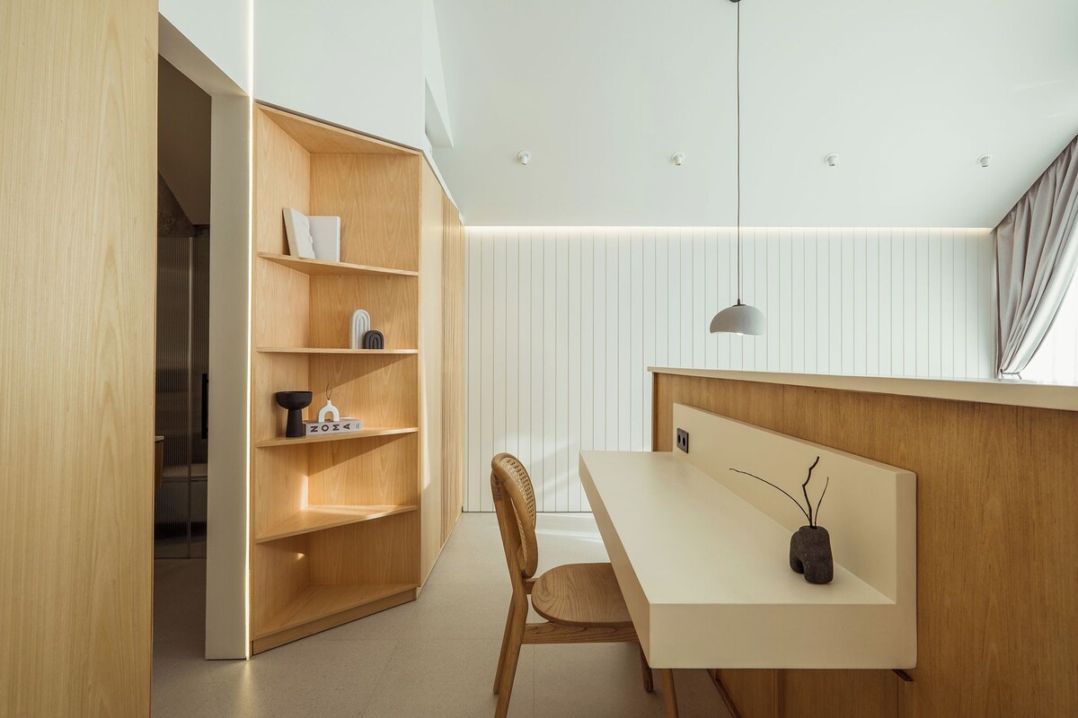 A modern, minimalist room with a wooden bookshelf, a dining table with a vase, and a hanging pendant light.
