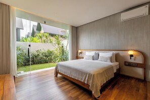 A modern bedroom with a large bed, wooden flooring, a glass door leading to a garden, and a built-in air conditioning unit.