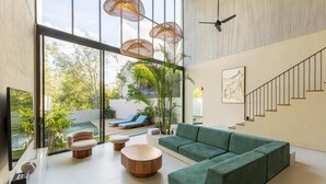 A modern living room with a green sofa, round wooden coffee table, and a TV. There are wicker lamps, a staircase, and a view of a pool and greenery outside.