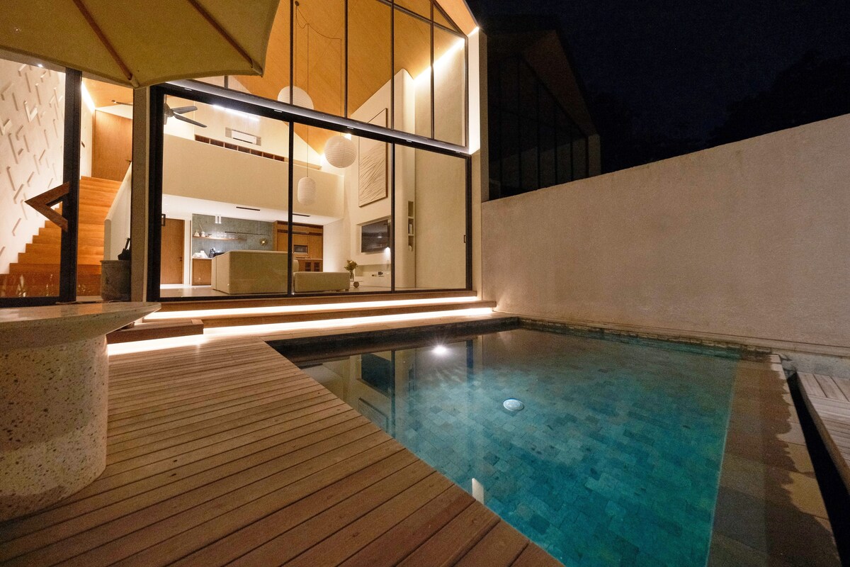 A modern outdoor swimming pool with a wooden deck, a glass-enclosed kitchen area, and a staircase leading to an upper level.