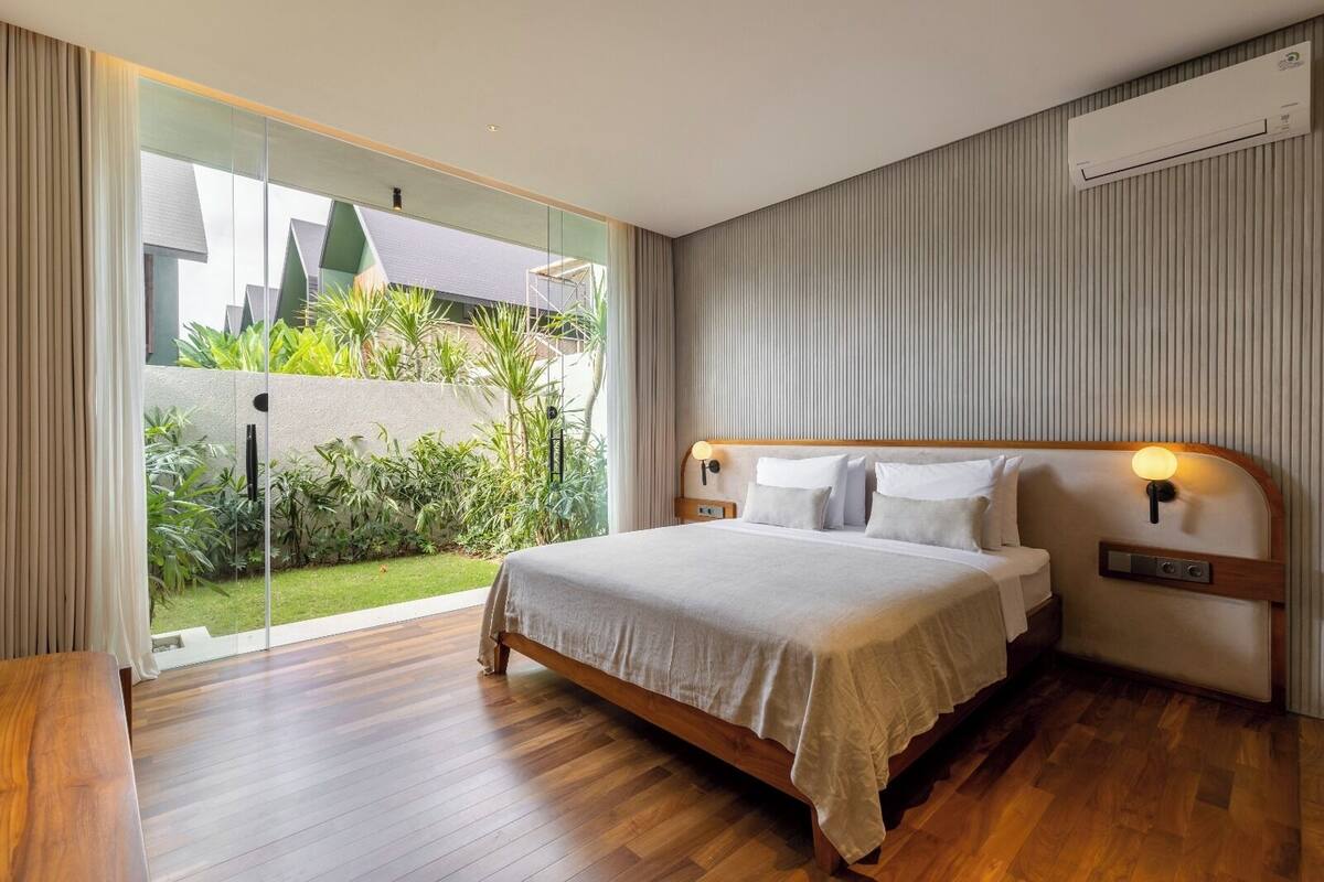 A modern bedroom with a large bed, wooden flooring, a glass door leading to a garden, and a built-in air conditioning unit.