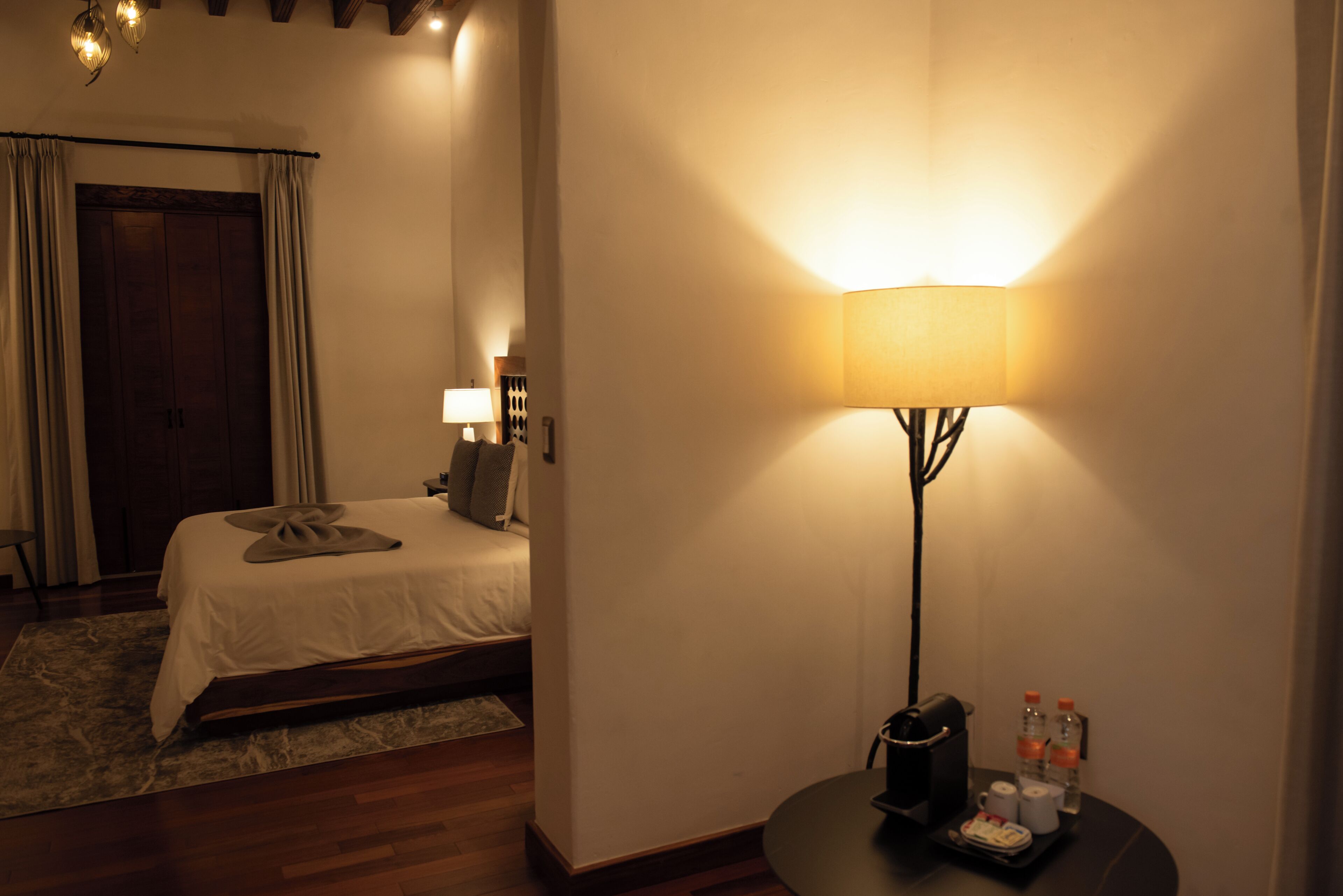Una habitación de hotel con una cama, una mesita de noche con una lámpara, una pequeña mesa con un neceser y una puerta.