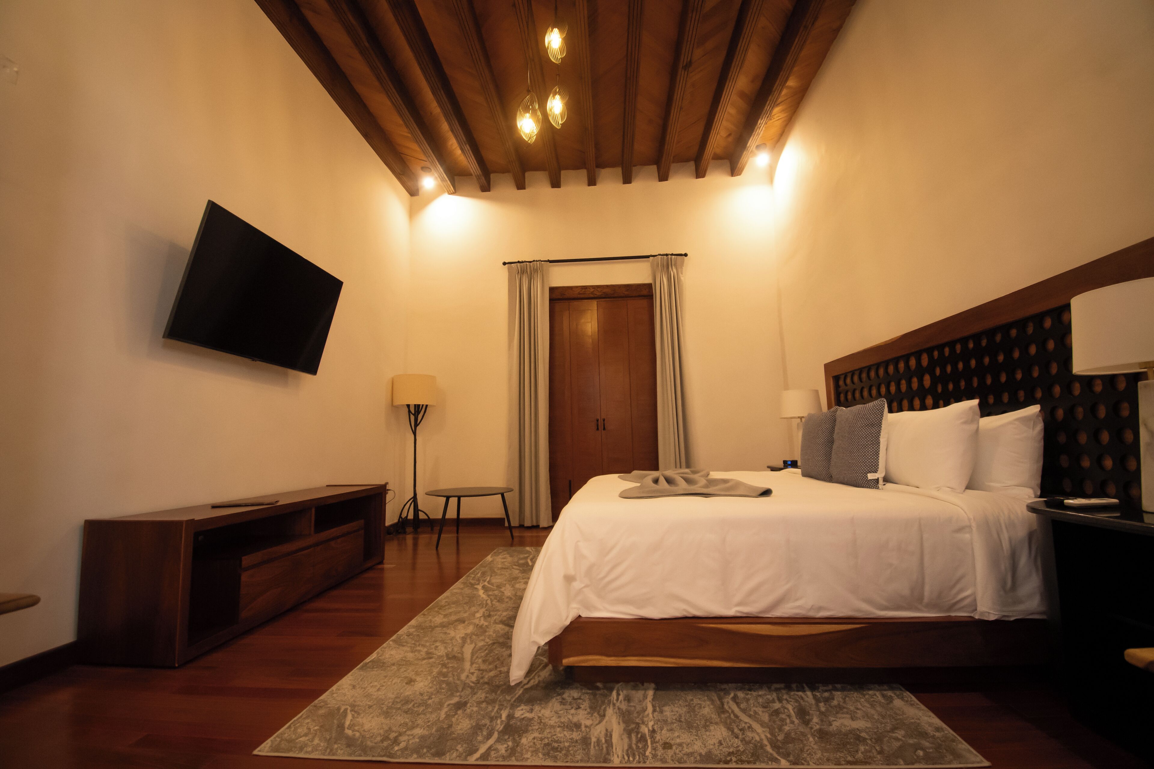 Un dormitorio con cama, televisor montado en la pared, cabecera de madera y una puerta con cortinas.