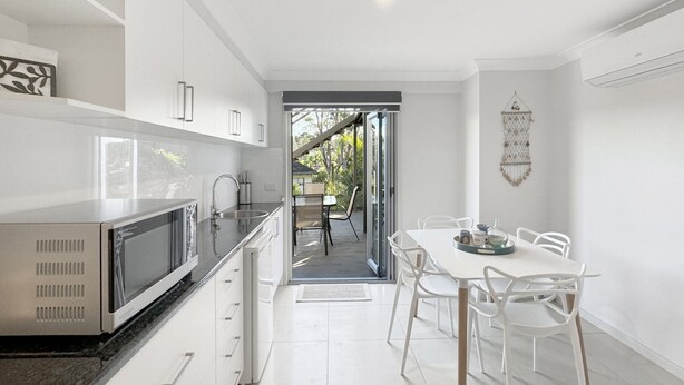 A modern kitchen with white cabinets, a stainless steel microwave, and a dining area with white chairs and a table. - Spacious Beach Studio With Alfresco Dining (Surf Beach)