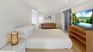 A bedroom with a bed, a brown sofa, a TV, and a sliding glass door. - Spacious Beach Studio With Alfresco Dining (Surf Beach)