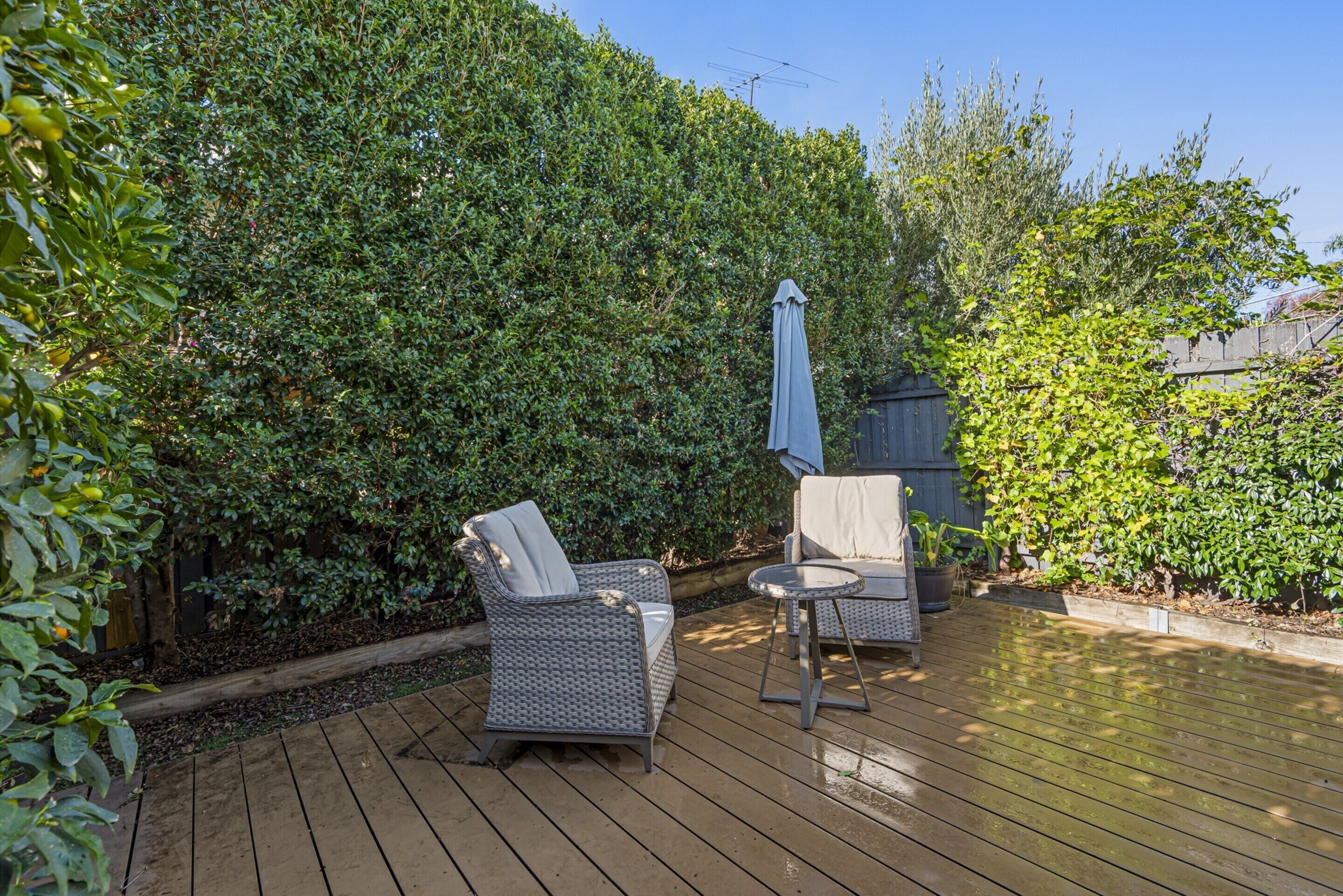 A wooden deck with wicker furniture, including two chairs and a small table, surrounded by dense greenery and a wooden fence.