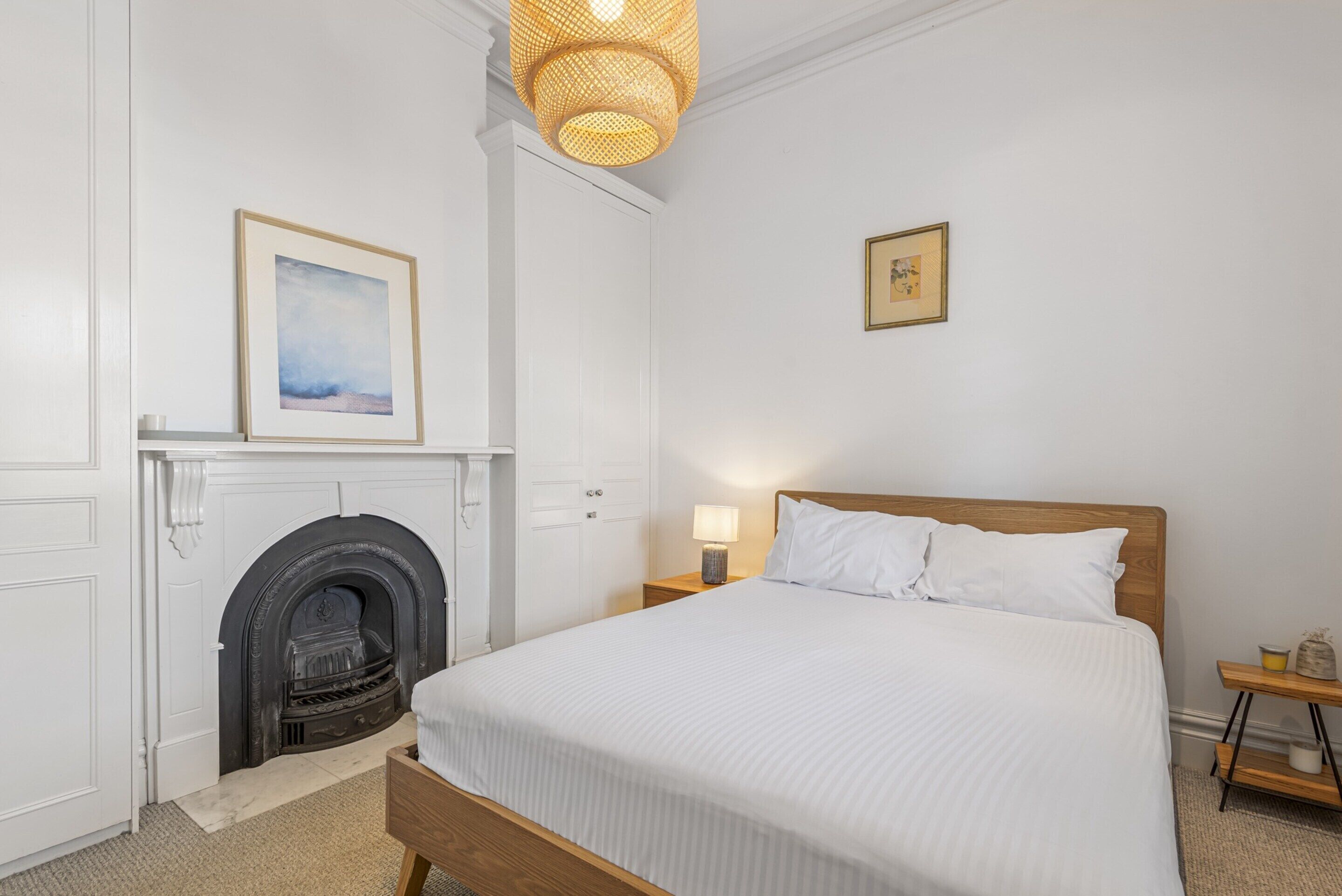 A bedroom with a bed, a fireplace, a nightstand with a lamp, and a framed picture on the wall.