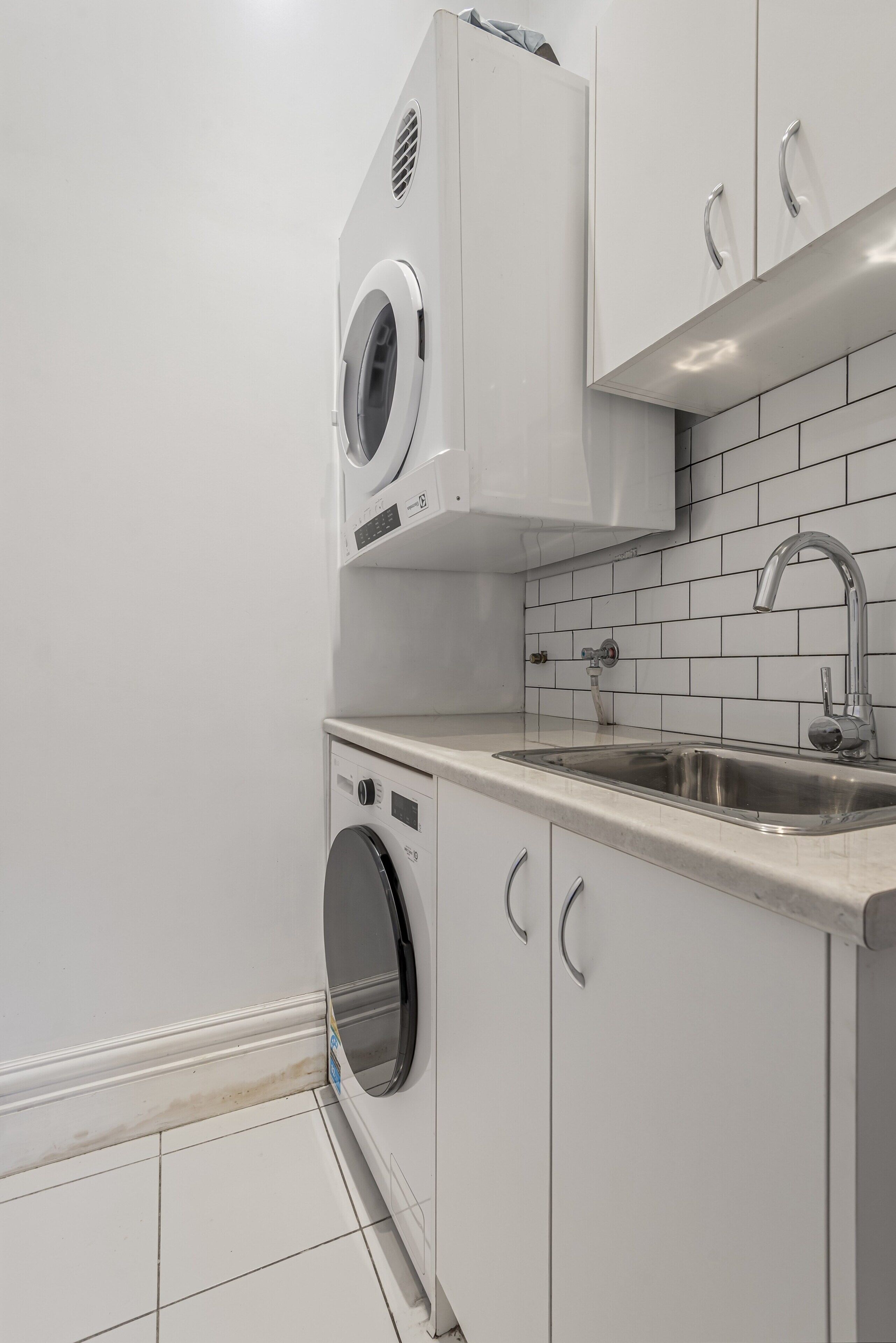 Une cuisine compacte dotée d’un lave-linge et d’un sèche-linge intégrés, d’un évier en acier inoxydable et d’un dosseret de carrelage blanc.