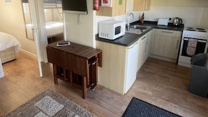 Una cocina compacta con microondas, pileta y cocina. Hay una pequeña mesa de madera y una cama en la habitación contigua.