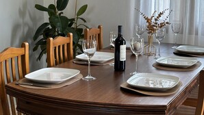 Mesa de comedor dispuesta con platos, vasos y una botella, rodeada de sillas de madera y una planta en maceta.