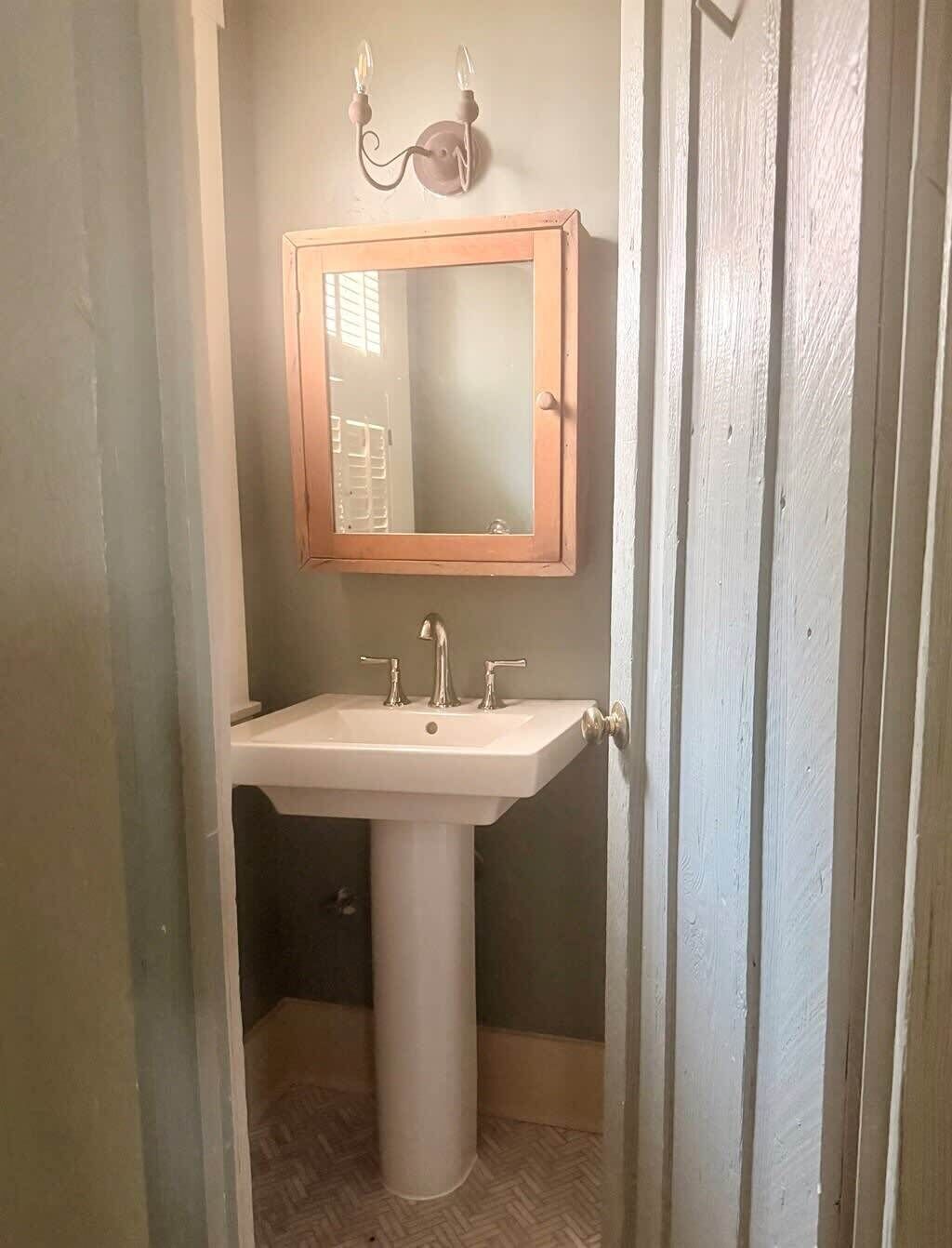 A bathroom with a white pedestal sink, a wooden-framed mirror, and a wall-mounted light fixture.