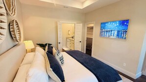 Quarto com cama, almofadas, mesa de cabeceira, abajur, pintura e porta para um banheiro.