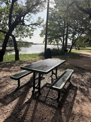 A picnic table with benches in a park with trees and a body of water in the background. - New Waterfront Home: Enjoy Lake LBJ Water Views From Every Bedroom | Luxury (Granite Shoals)