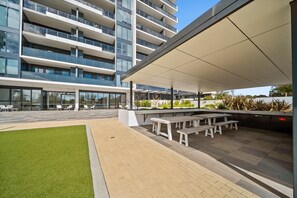 A modern outdoor area with a covered seating area, a grassy lawn, and a multi-story building in the background. - Stylish 2-bed Apartment With Luxe Amenities Access (Canberra)