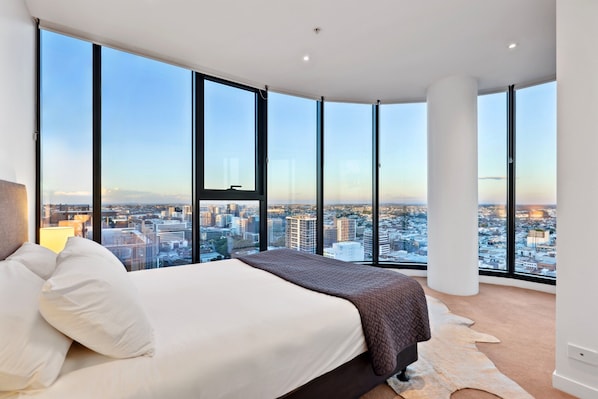 Une chambre moderne avec un grand lit, une vue sur la ville grâce à de grandes fenêtres du sol au plafond et un tapis au design original.