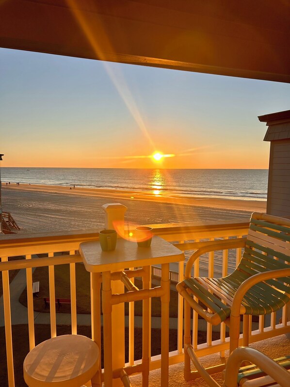 A balcony with a view of the sunset over the ocean, a table with a candle, and a chair.