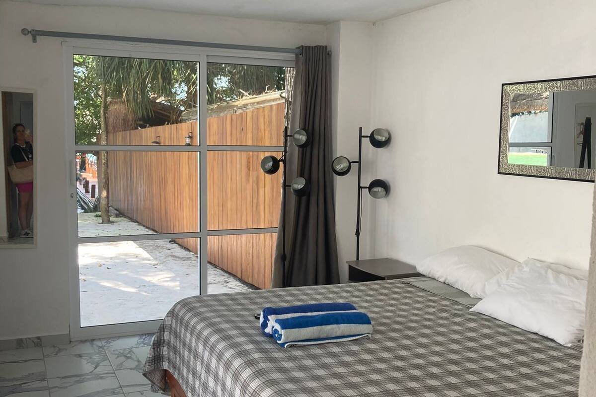 A bedroom with a bed, a large window, a mirror, and a view of the outside.