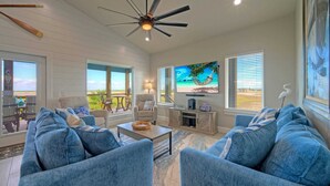 A spacious living room with a large TV, comfortable seating, and a view of the outdoors. - Bayfront Home w/ Pool, Game Room + Walk to Beach & Golf Cart Access Pelicans Perch by Avantstay (Port Aransas)