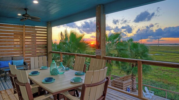 A covered outdoor dining area with a table set for four, chairs, and a view of the sunset over a grassy landscape. - Bayfront Home w/ Pool, Game Room + Walk to Beach & Golf Cart Access Pelicans Perch by Avantstay (Port Aransas)