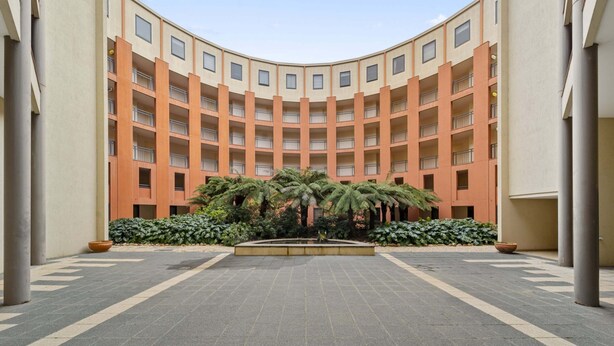A courtyard with a central fountain, surrounded by palm trees and a multi-story building with balconies. - Loft-style CBD 1-bed With Rooftop Terrace & BBQ (Canberra)