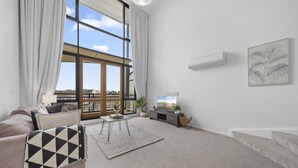 A modern living room with a large window, a sofa, a coffee table, and a TV stand. - Loft-style CBD 1-bed With Rooftop Terrace & BBQ (Canberra)
