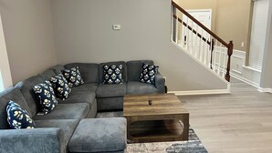 Un salon moderne avec un canapé d’angle, un pouf et une table basse. On aperçoit un escalier avec une rampe en bois à l’arrière-plan.