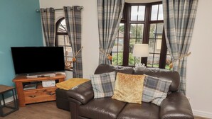 A living room with a brown leather sofa, a wooden TV stand with a flat-screen TV, and plaid curtains. - Nobles Nook (Seahouses)