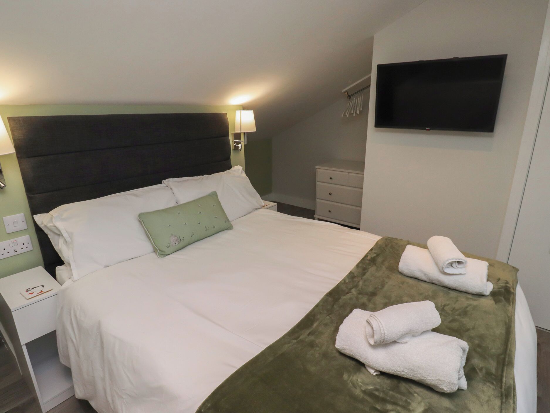 Un lit bien fait, avec du linge de lit blanc et un coussin vert, un plaid vert et deux serviettes enroulées posées sur le lit.