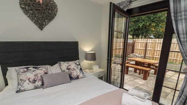 A bedroom with a bed, a bedside table with a lamp, a breakfast tray with croissants and orange juice, and a view of an outdoor patio with a table and chairs. - Nobles Nook (Seahouses)