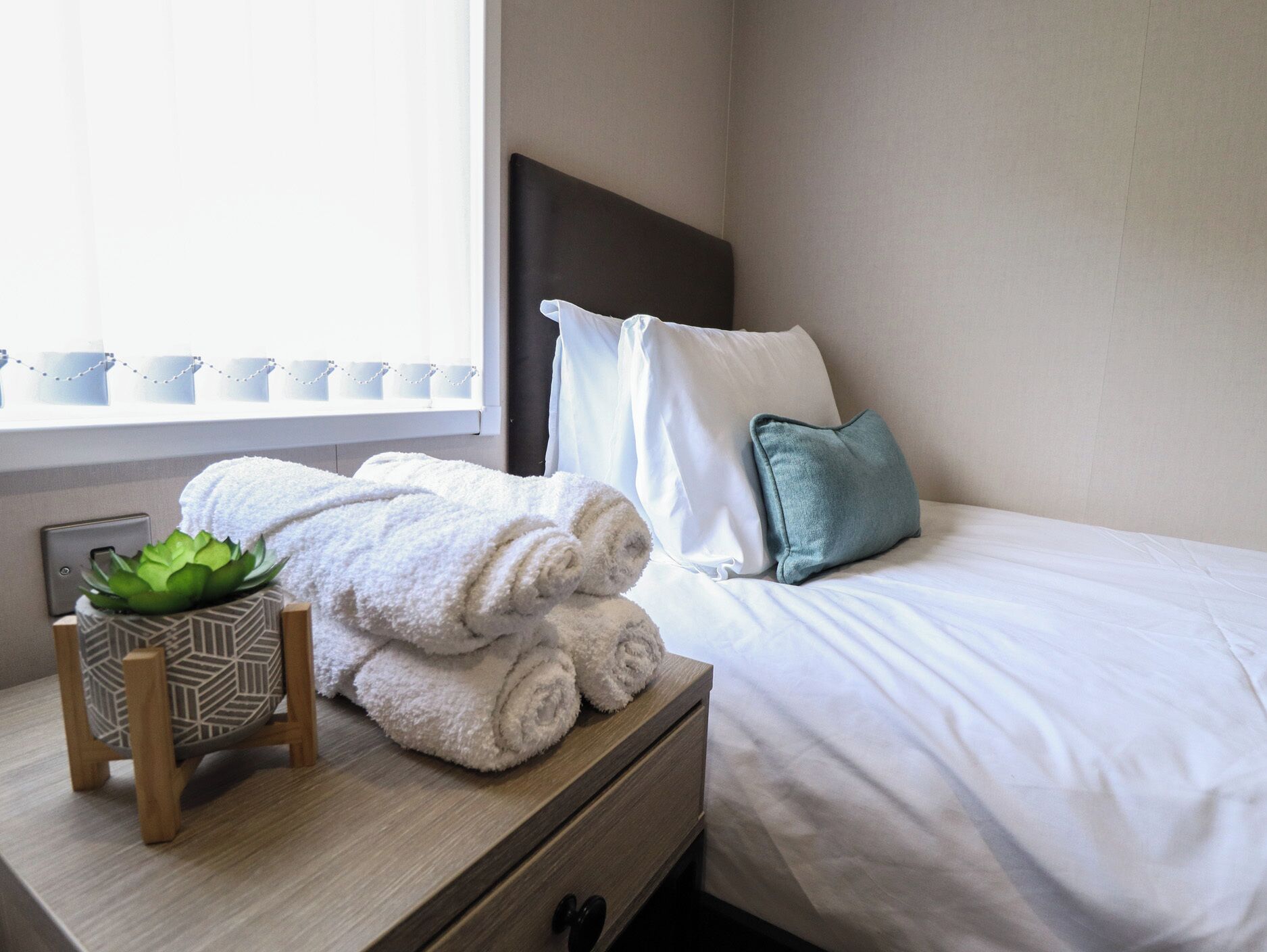 Een net opgemaakt bed met witte lakens, een teal kussen en opgerolde witte handdoeken op een houten nachtkastje naast een raam.