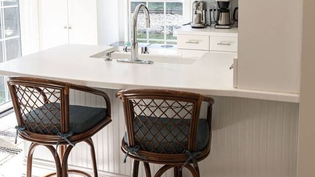 Una cocina con muebles blancos, un ventanal grande y dos taburetes de mimbre.