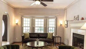 Una sala de estar con un sofá de cuero oscuro, dos sillones verdes, una mesa de centro de vidrio y una chimenea.