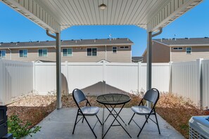 Un patio techado con dos sillas y una mesa, una cerca blanca y un edificio al fondo.