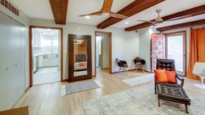 Uma sala de estar espaçosa com piso de madeira, um ventilador de teto, uma poltrona de couro com almofadas e vista para a cozinha através de uma porta aberta.