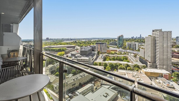 A balcony with a table and chairs, overlooking a cityscape with various buildings and greenery. - Elegant 2-bed Apartment With Balcony & City Views (Melbourne)