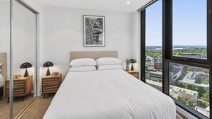 A modern bedroom with a large bed, a city view, a framed artwork on the wall, and a nightstand with a lamp and phone. - Elegant 2-bed Apartment With Balcony & City Views (Melbourne)