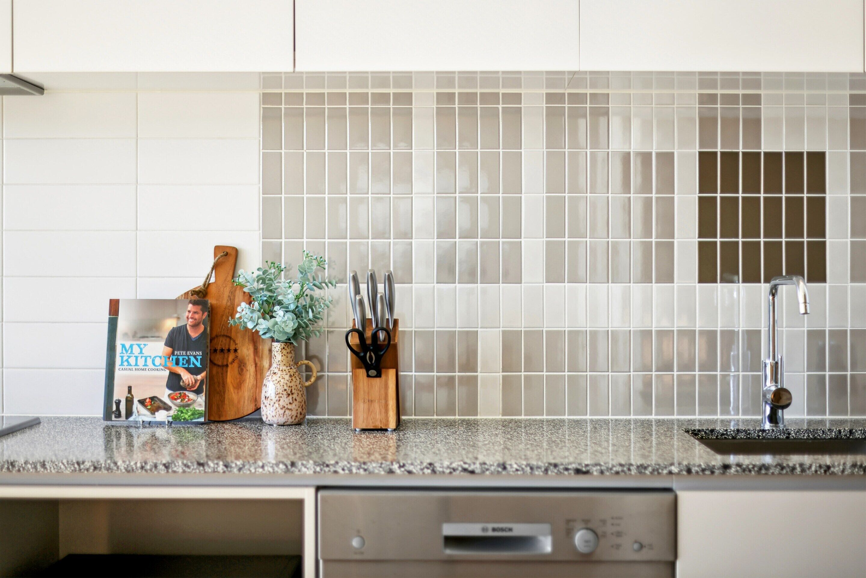 A kitchen with a Bosch dishwasher, a tiled backsplash, and a countertop with a book, a plant, and a knife block.