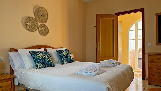 A neatly made bed with white bedding and tropical leaf-patterned pillows, a wooden nightstand with a drawer, and a wooden dresser with drawers. - Casa Luz de Azahar - Ole Solutions (Jerez de la Frontera)