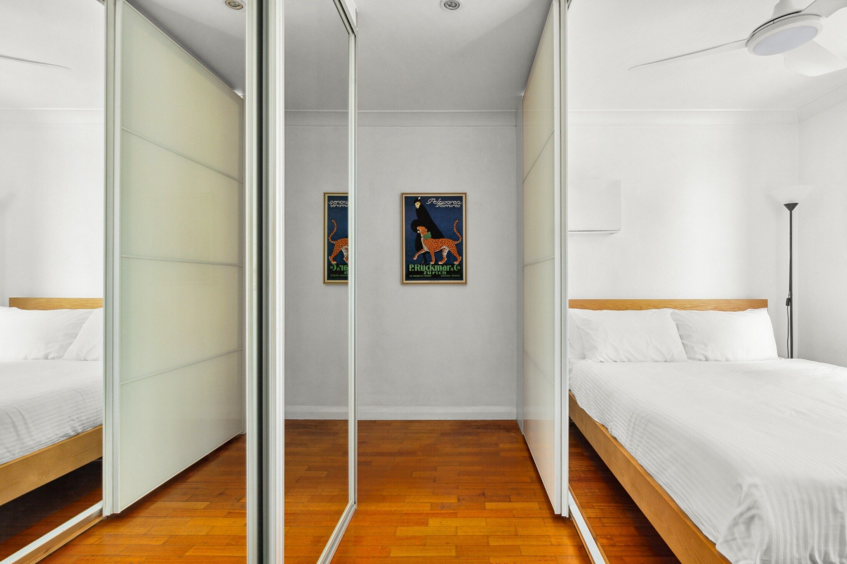 A modern hotel room with two single beds, a wooden floor, a ceiling fan, and two posters on the wall.