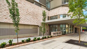 A modern building with a brick facade, a glass entrance, and a small garden area with young trees. - Bright 2-bed Apartment With Gym, BBQ & Pool Access (Canberra)