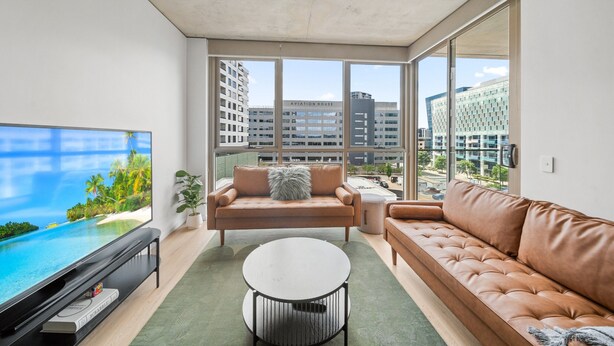 A modern living room with a large TV, a sofa, a coffee table, and a view of the cityscape through large windows. - Bright 2-bed Apartment With Gym, BBQ & Pool Access (Canberra)