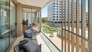 A balcony with a glass door, a seating area with cushions, and a view of a high-rise building and a green space. - Bright 2-bed Apartment With Gym, BBQ & Pool Access (Canberra)