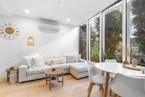 A modern living room with a sectional sofa, a round dining table with chairs, a wall clock, and large windows offering a view of greenery outside. - Brighton 1-bed Apartment With Balcony & Parking (Melbourne)