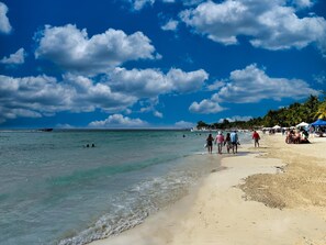 A beach with clear turquoise water, people walking along the shore, and a boat in the distance.