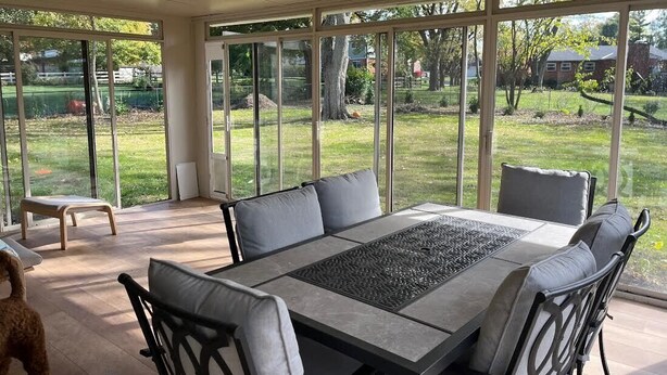 Un patio ensoleillé avec une table et des chaises, de grandes fenêtres offrant une vue sur une pelouse et des arbres.