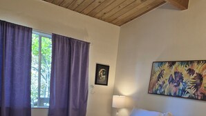 Een slaapkamer met een groot bed, nachtkastje, lamp, schilderij en een raam met gordijnen.