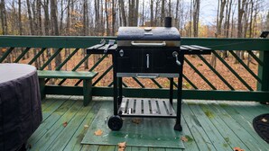 Una parrilla portátil sobre una terraza de madera con barandilla verde, rodeada de árboles.